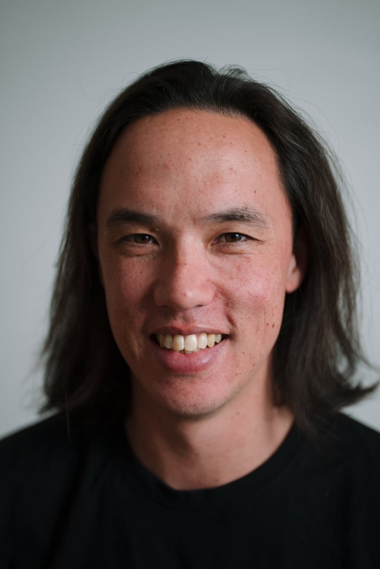 Portrait of a smiling man with long dark hair wearing a black shirt against a light gray background
